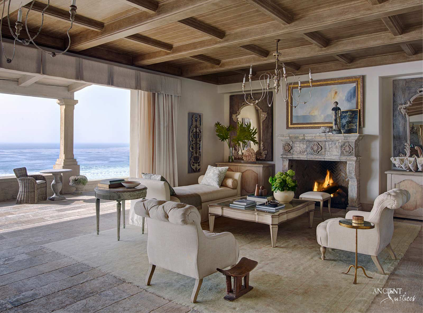 Ocean view living room with a carved stone fireplace mantel bearing quatrefoil rosettes, weathered grey limestone plank floors and an open loggia with a stone column facing the sea