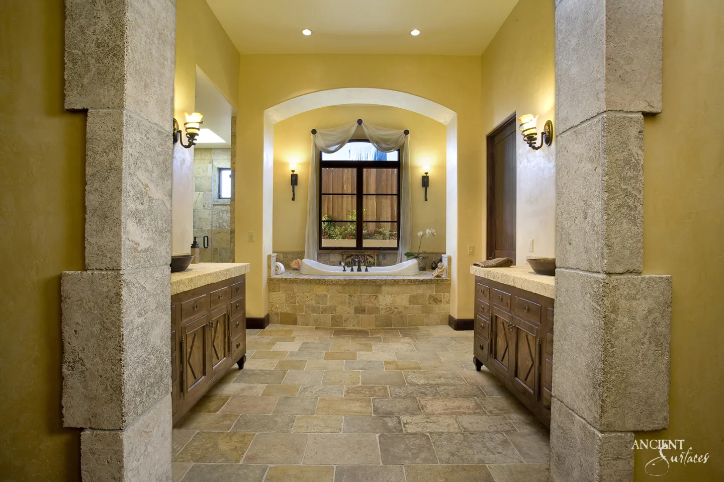 Bathroom seen through a portal of large textured limestone blocks, with a tumbled limestone floor in grey and ochre, two vanities with thick stone tops and a stone clad tub in an arched alcove