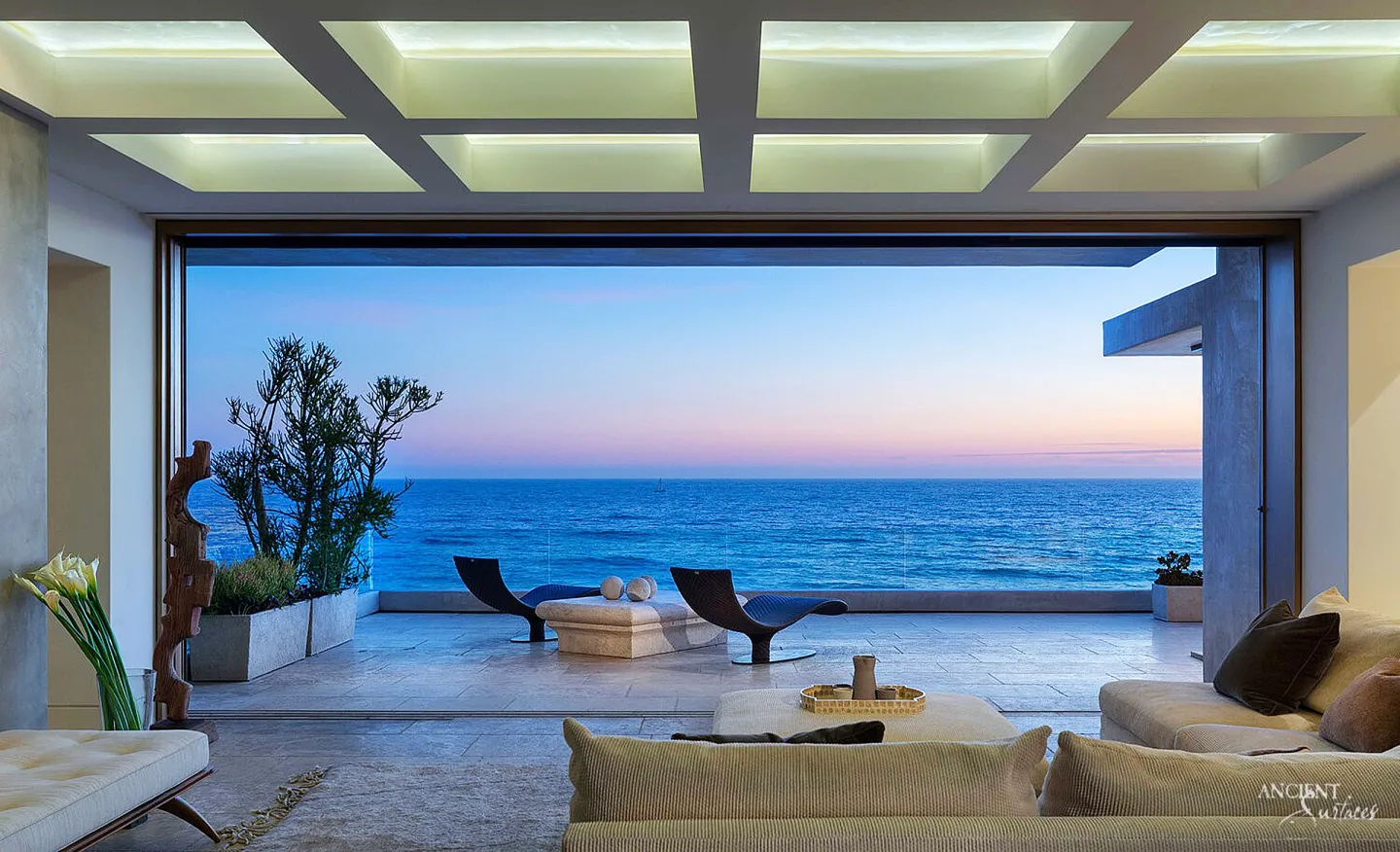 Contemporary living room under a coffered ceiling opening fully onto an ocean terrace paved in pale limestone planks, with two loungers beside a low stone block table