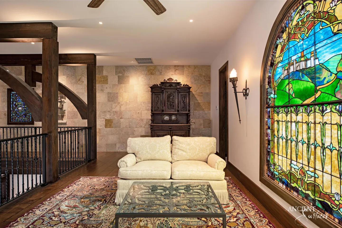 Upper landing with a wall clad in squared cream, honey and grey limestone blocks behind a carved wooden cabinet and sofa, timber gallery posts and a large arched stained glass window
