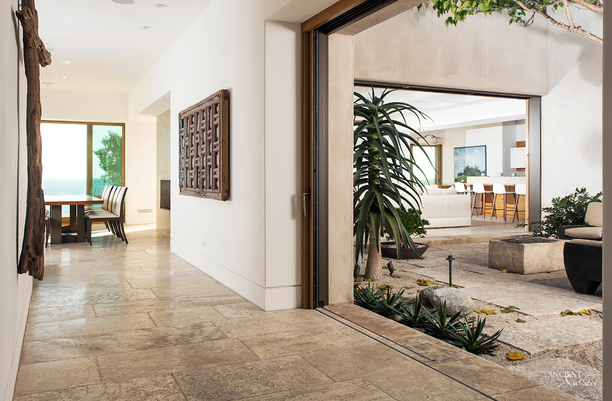 Hallway paved in long pale limestone planks of random length leading to an ocean view dining room, beside an open pocket door to a courtyard with stone pavers, gravel and aloes