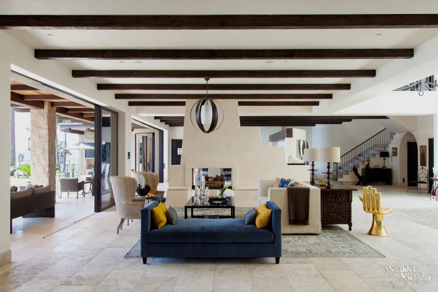 Long open plan living room with a pale cream limestone floor running past a plastered double sided fireplace to the stair hall and out through sliding doors to a covered loggia