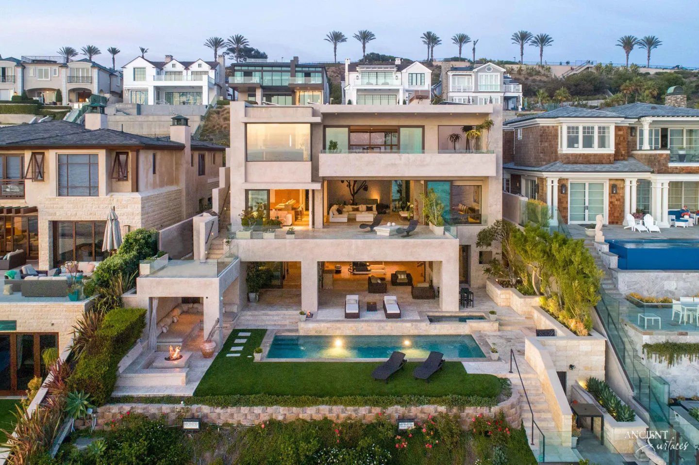 Aerial view at dusk of a contemporary coastal villa with pale limestone terraces, pool and spa surrounds, broad stone steps and stepped garden walls of squared stone blocks