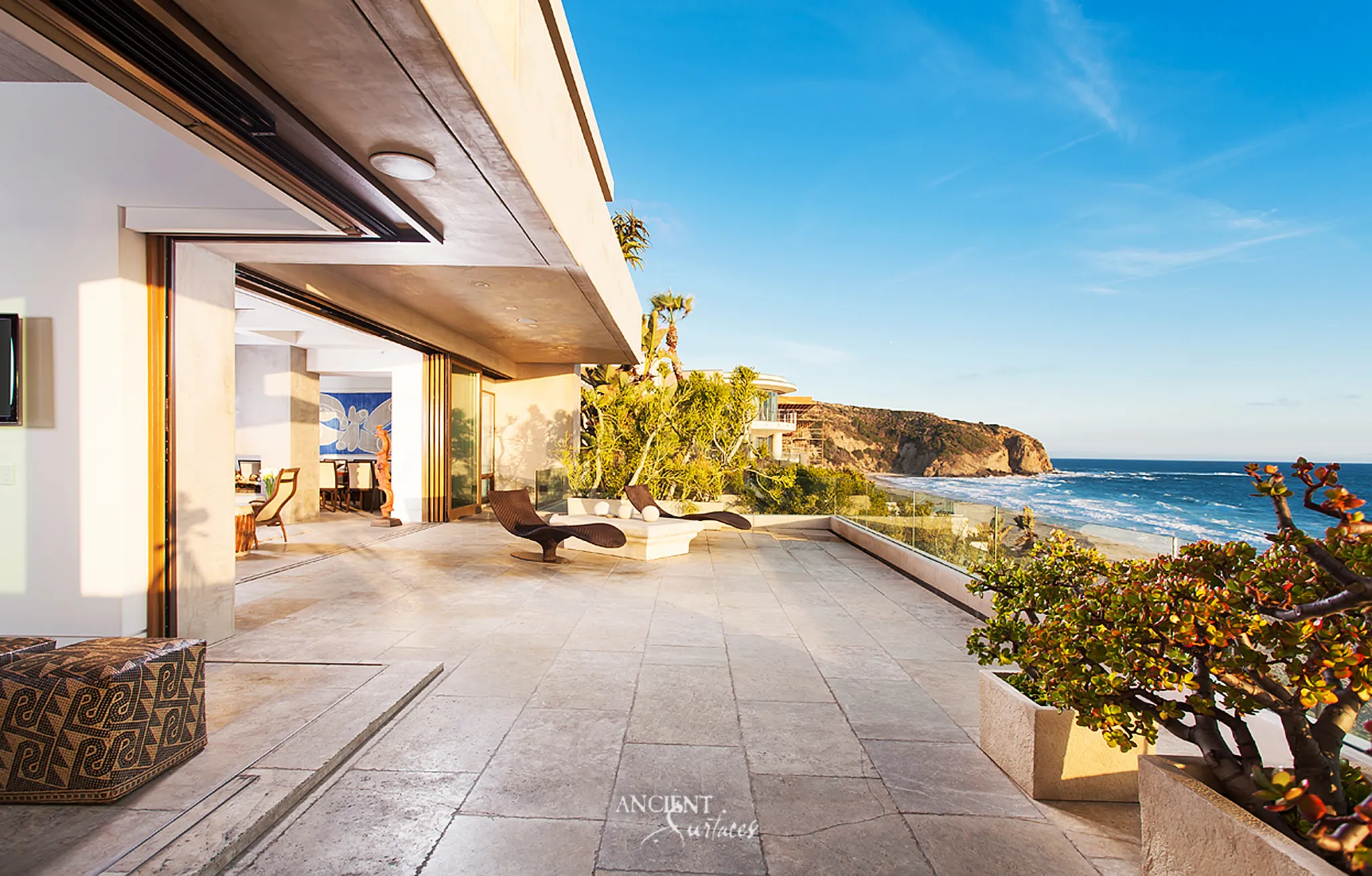 Ocean terrace paved in large pale limestone slabs continuing through opened glass walls into the living and dining rooms, with stone planters along a glass balustrade and a view of the coastline